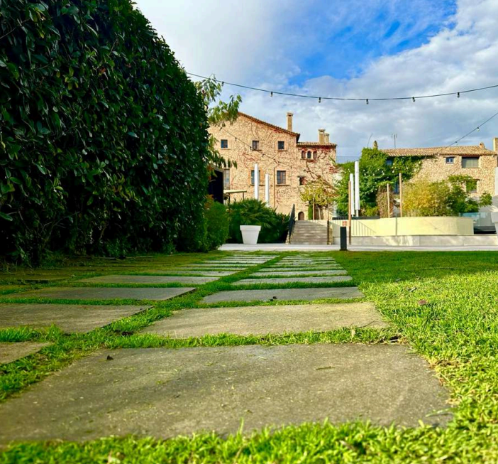 Sendero de piedras en un jardín verde, con un seto a la izquierda y edificios de piedra al fondo. El cielo está parcialmente nublado y hay luces colgantes en la parte superior.