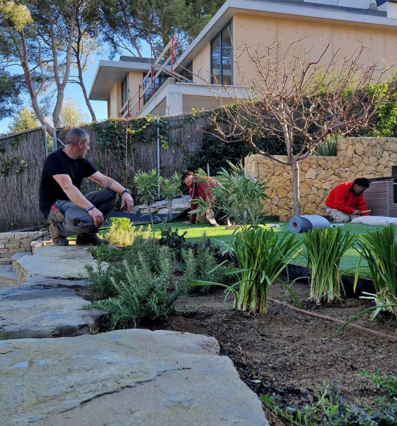 Tres personas trabajando en un jardín frente a una casa moderna. Están instalando césped y plantando vegetación. Uno de ellos está arrodillado mientras los otros dos están trabajando con herramientas.