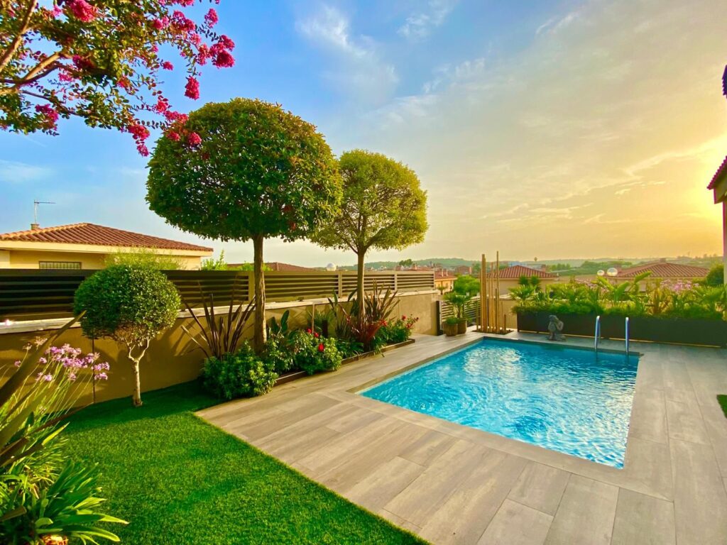 Jardín con una piscina pequeña y bien cuidada rodeada de plantas y árboles. Al fondo, un atardecer con cielo anaranjado y casas.