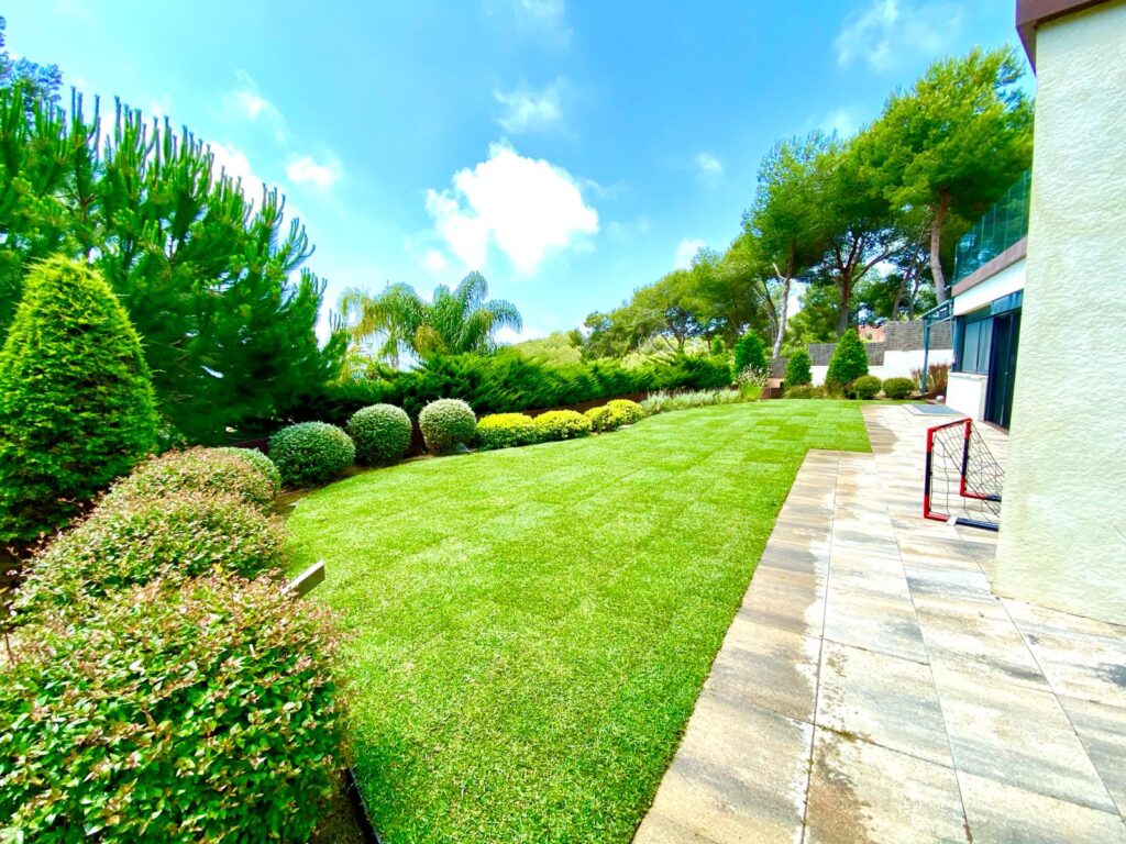 Amplio jardín con césped verde bien cuidado, arbustos podados y árboles. A la derecha, una terraza pavimentada.