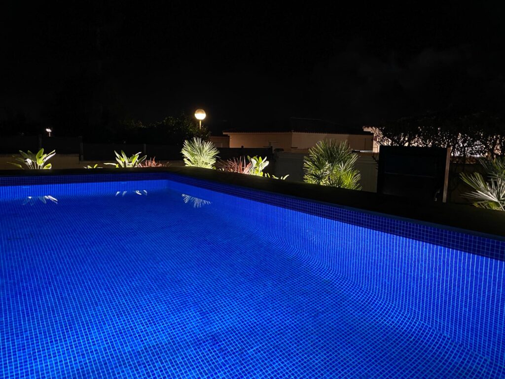 Piscina iluminada de noche con luces azules y plantas alrededor. Se pueden ver luces de casas al fondo.