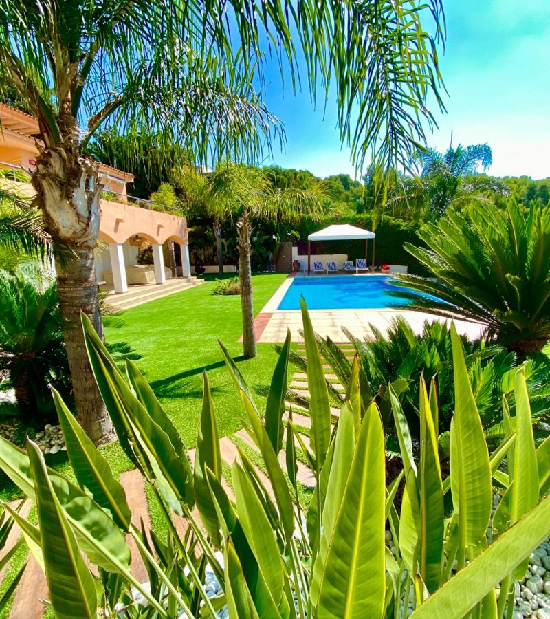 Jardín tropical con palmeras y plantas exóticas alrededor de una piscina azul. A la izquierda, un edificio con arcadas y un césped bien cuidado.